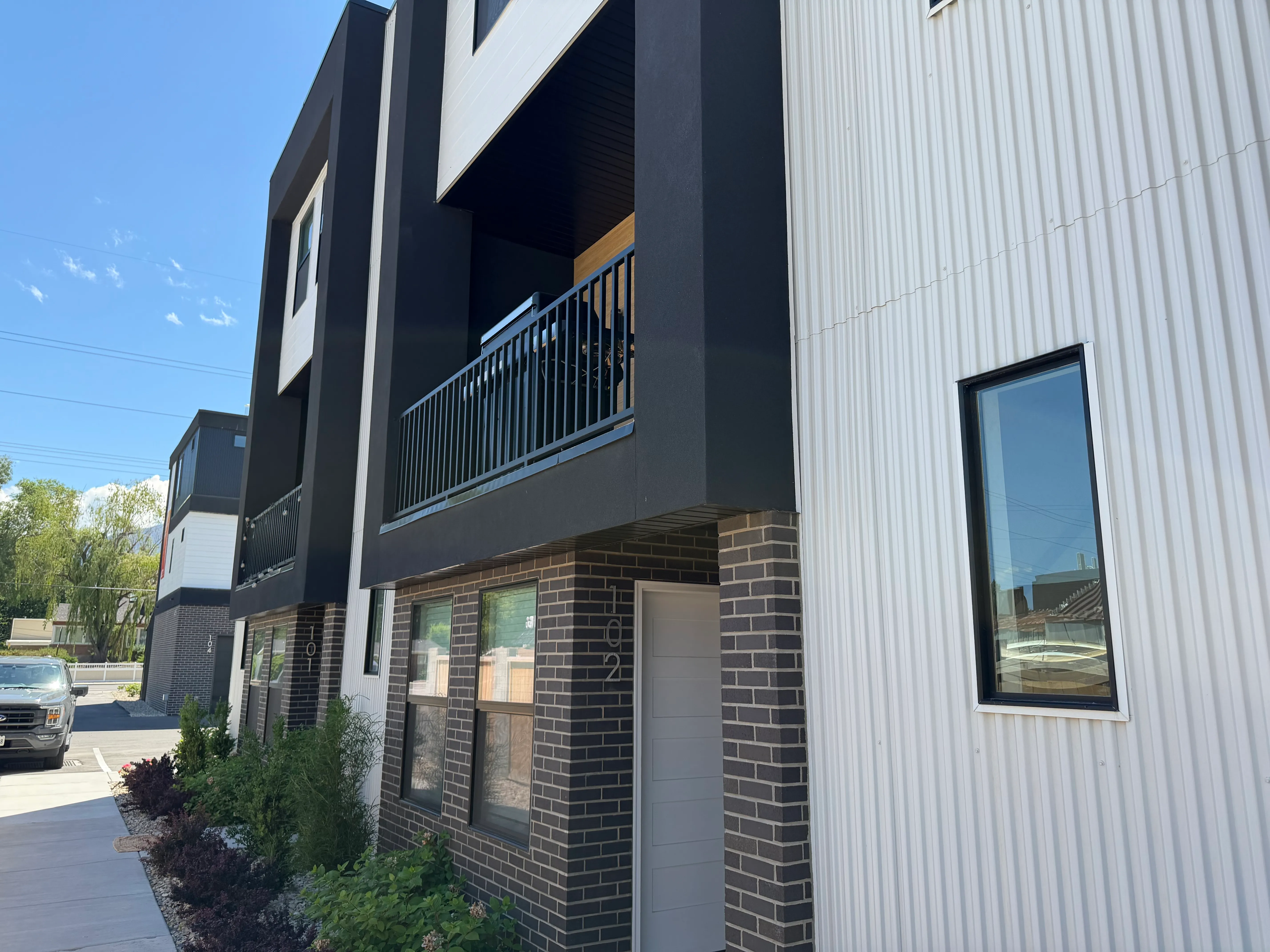 Side view of the Quincy townhomes with modern black-and-white siding, private balconies, and a brick base under a clear blue sky